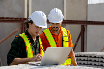 Two civil engineers are using computer notebook and tablet to monitor precast production progress in a factory. Construction managers are using technology to improve their factory system
