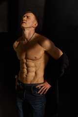 Fototapeta premium Slim muscular male model on black background. Shirtless fitness guy in black sweatpants posing in studio. Man with six pack abs in sunset light. Brutal photo