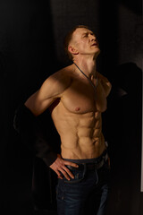 Fototapeta premium Slim muscular male model on black background. Shirtless fitness guy in black sweatpants posing in studio. Man with six pack abs in sunset light. Brutal photo