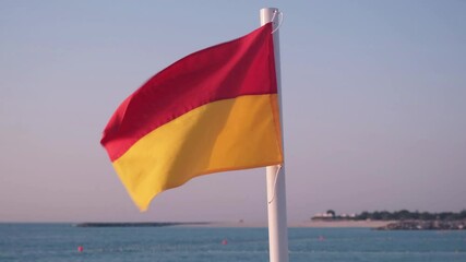 Red and yellow beach safety flag on a pole fluttering against a clear blue sky by the sea. Lifeguard signal indicating swimming safety conditions at a coastal area.