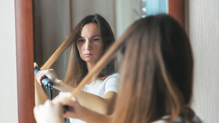 Young woman using a hair iron and doing a hairstyle looking at the mirror in the morning at bedroom...