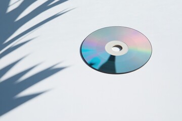 polished cd disc laying flat on empty white surface encircled by delicate play of light and shadows which dance around