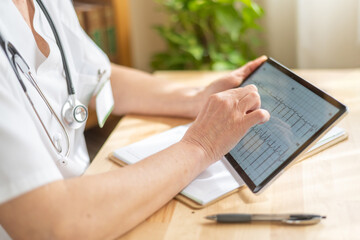 Senior female doctor wearing a stethoscope sits at her desk, using a digital tablet to analyze...