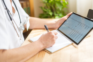 Cardiologist examining electrocardiogram data on a digital tablet and writing observations on a notepad during a medical consultation, focusing on healthcare and telemedicine