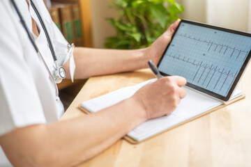 Doctor wearing stethoscope is using a digital tablet displaying an electrocardiogram while taking notes on a medical report during a consultation in a medical office