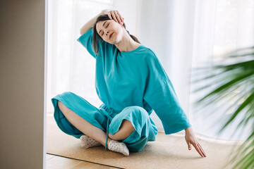 Young white woman in a turquoise robe does a neck stretch while sitting cross-legged on a mat in a...