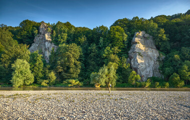 Rear view of a bearded traveler with a backpack standing on a rocky riverbank, admiring the scenic view of lush green mountains and towering rock formations.