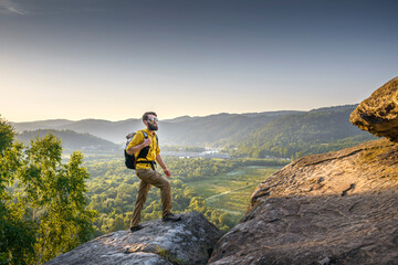 Obraz premium Bearded male hiker in a yellow shirt standing on a rocky cliff at sunset, overlooking a scenic valley with river, forest, and distant mountains.