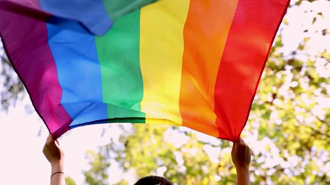 Slow motion video of a woman raising a vibrant rainbow pride flag outdoors, with sunlight filtering through trees in the background. The flag symbolizes LGBTQ+ pride and inclusivity