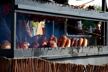 A close up on a massive outdoor grill with numerous sizzling tipes of meat, including steaks, sausages, bacon, and ham, sold to public during a medieval fair in Poland on a warm summer day
