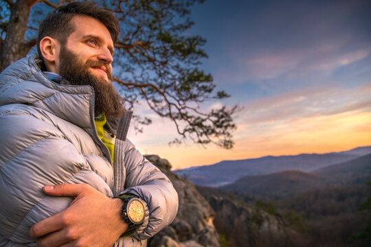 Bearded man in a puffer jacket stands on a mountain at sunset, arms crossed, wearing a Casio watch. He smiles confidently, with rocky peaks, autumn forests, with a focus on his travel wristwatch	