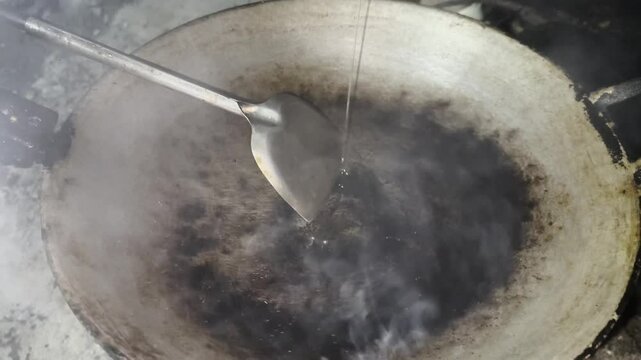 Asian cook is cooking in thai kitchen in local traditional country style, preparing  streaming vegetable oil being poured into a hot traditional wok to cooking high heat stir frying food