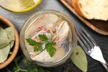 Pieces of delicious herring with spices in jar and bread on black table, flat lay