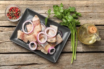 Pieces of herring with onions, peppercorns, oil and parsley on wooden table, flat lay