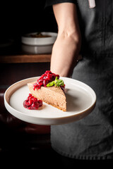 A slice of cheesecake topped with berry compote and a mint leaf is presented on a white plate by a person wearing a dark apron. The background is blurred, focusing attention on the dessert.
