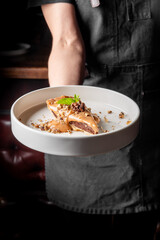 A person in a dark apron holds a white plate with a dessert featuring a chocolate base, caramel topping, nuts, and a mint garnish.
