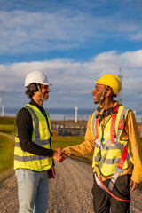 Two multi ethnic engineers shaking hands in front of a wind power station, celebrating successful teamwork and sustainable energy development