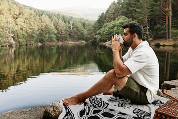Young man drinking coffee at a picnic by a lake with a mountain and forest landscape