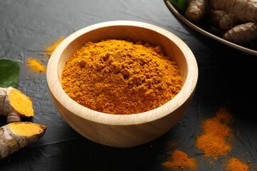 Aromatic turmeric powder in bowl and fresh roots on black table, closeup