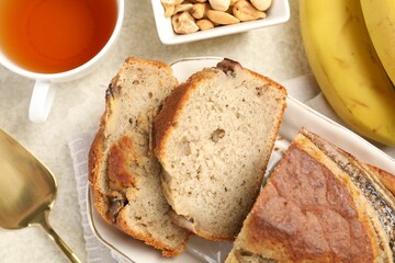 Cut banana bread with nuts, fruits, server and tea on light grey table, flat lay
