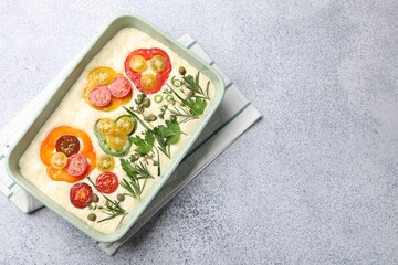Raw focaccia with vegetables and spices in baking dish on light grey table, top view. Space for text