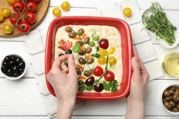 Woman adding basil to raw focaccia at white wooden table, top view