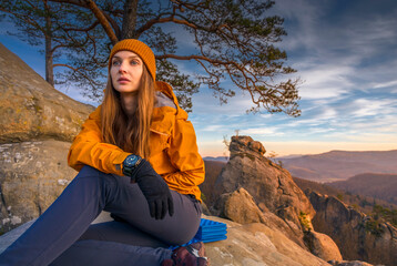 Young woman in an orange jacket and beanie sits on a mountain peak at sunset, wearing a Casio watch...