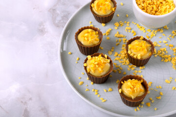 Chocolate cups with open mango filling and orange crunch sprinkles on a ceramic plate on light background. Holiday dessert. Handmade sweets