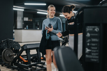 Two women in fitness attire share a laugh and chat while working out in a modern gym setting surrounded by gym equipment.