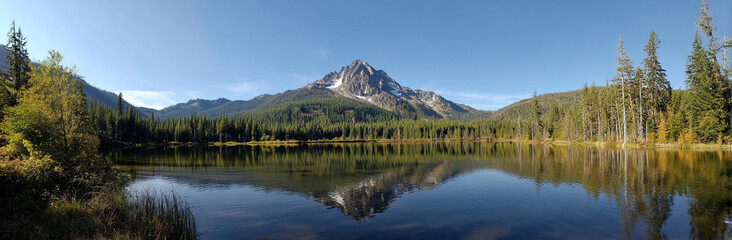 Panoramic photo of Lake. The lake is clear and calm, reflecting the surrounding mountains and trees in its surface. In front of it stands Mount Hood with snow-capped peaks against a blue sky backgroun
