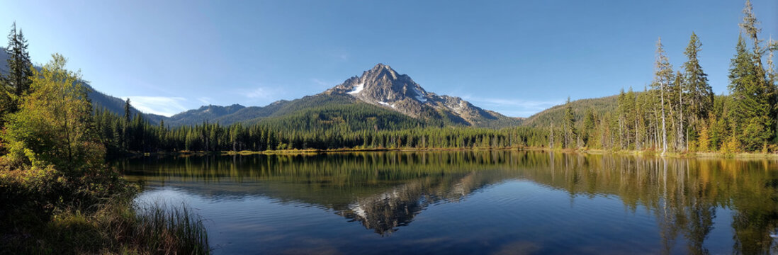 Panoramic photo of Lake. The lake is clear and calm, reflecting the surrounding mountains and trees in its surface. In front of it stands Mount Hood with snow-capped peaks against a blue sky backgroun - Powered by Adobe