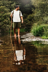 Hiker crossing a stream barefoot in a green forest with a walking stick and backpack