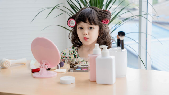 Cute little girl with hair curlers playing with makeup at vanity table, making a kissy face in front of a mirror, imitating adult beauty routine playfully.
