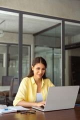 Focused smiling latin female marketing manager, professional ai specialist working at laptop computer sitting at desk in modern office. Young woman student using pc for online business studying course