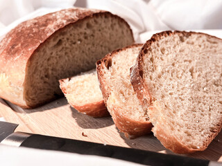 Freshly baked round bread cut into slices on a wooden board with a knife on a white fabric background. Close up.