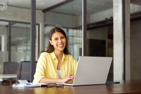 Cheerful young woman employee using pc for business ai work. Portrait at camera smiling latin female studying student, marketing specialist working at laptop computer sitting at desk in modern office