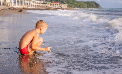 Fototapeta premium Little boy crouching on wet sand by the sea shore observing waves