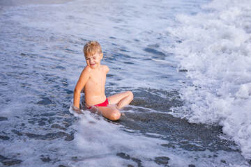 Fototapeta premium Little Boy in Swimwear Relaxing in the Foamy Shoreline Waters at the Beach