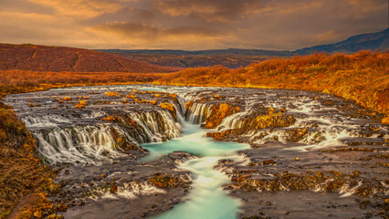 Brúarfoss Waterfall in Iceland glows beneath a golden sky. Its turquoise stream cuts through...