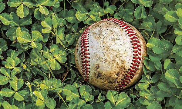 A baseball sits among lush green clover, aged and earthy, with intricate red stitching detail