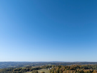 Panoramic view of a rural landscape under a clear blue sky, showcasing rolling hills, fields, and scattered houses surrounded by trees.