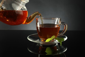 Pouring freshly brewed tea from teapot into glass cup on black table, closeup