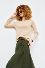 Fototapeta premium Stylish woman with curly hair in a beige sweater and green skirt, posing confidently against a plain background, emphasizing fashion and modern aesthetics in a minimalistic setting