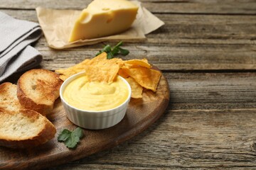 Tasty cheese dipping sauce in bowl, nacho chips and croutons on wooden table, closeup. Space for text