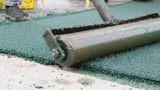A worker uses a cylindrical metal roller to smooth and compact freshly installed EPDM rubber granules. A compactor roller is in the process of leveling the floor covering on a new outdoor playground.