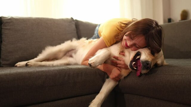 A cheerful teen girl is cuddling her golden retriever on a soft couch inside a bright living room. Sunlight streams in, creating a warm atmosphere as they enjoy each other's company.