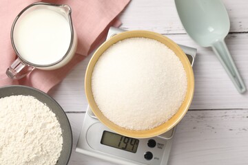 Digital kitchen scale with bowl of sugar and other products on white wooden table, flat lay