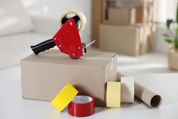 Fototapeta premium Dispenser with adhesive tape rolls and cardboard box on desk indoors