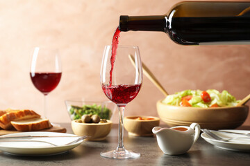 Pouring red wine into glass at grey table with tasty food, closeup