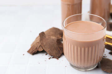 Tasty chocolate milk in glass and pieces of chocolate on white tiled table, closeup. Space for text